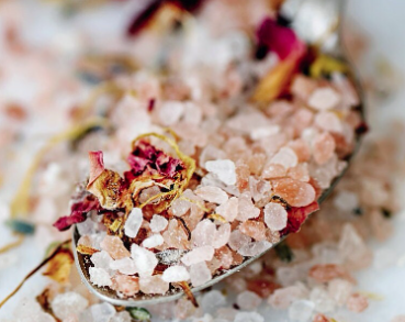 A spoonful of bath salts with pink and white crystals, garnished with petals and herbs, indicating a blend of botanical ingredients.